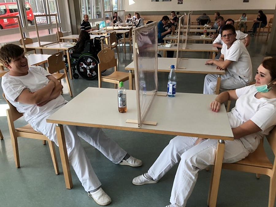 Wir blicken in den Speiseraum der Werkstatt auf rechteckige Tische auf denen in der Mitte eine Folientrennwand steht. An den Kopfseiten der Tische sitzen die Mitarbeiter.