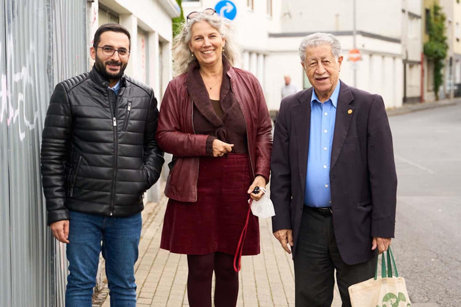 Eine Frau stehet zwischen zwei Männern. Alle drei schauen in die Kamera und lachen.