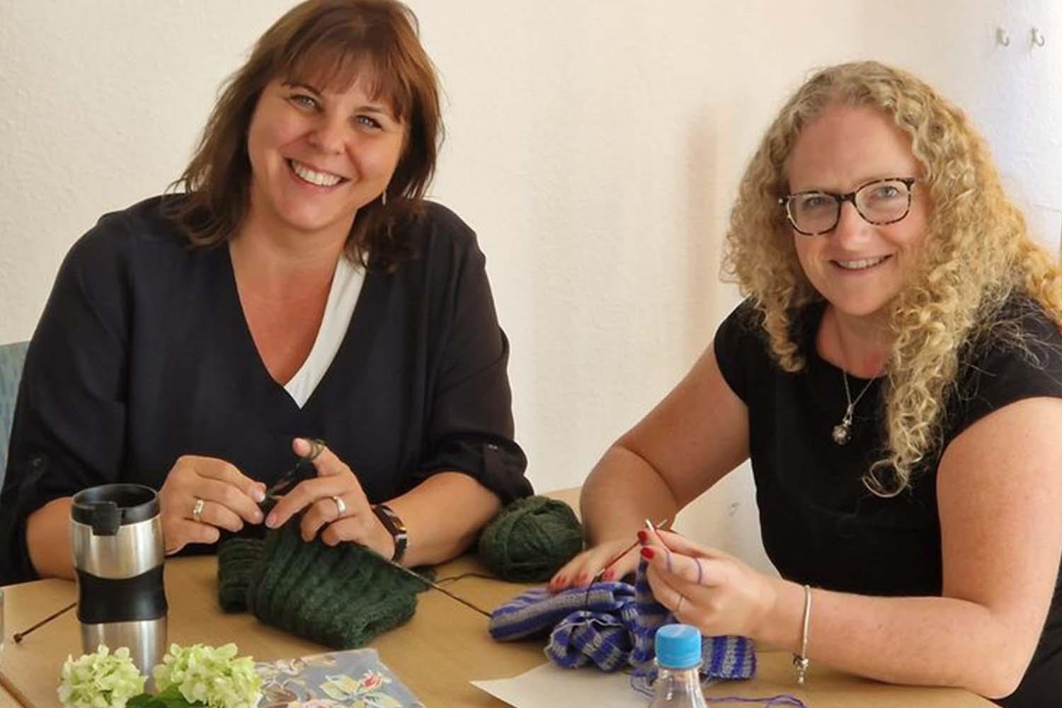 Zwei Frauen sitzen am Tisch, stricken und lachen.
