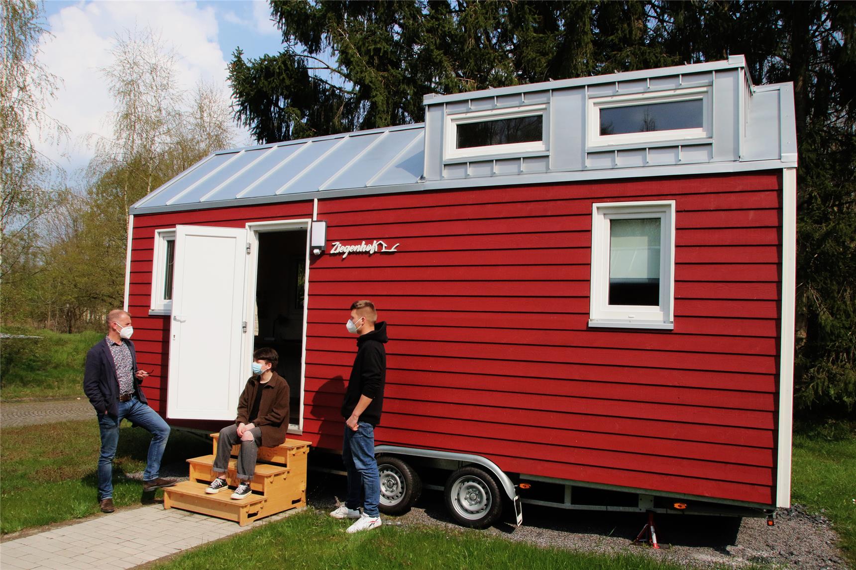 Das Foto zeigt drei Menschen vor einem Tiny House stehen und sitzend.